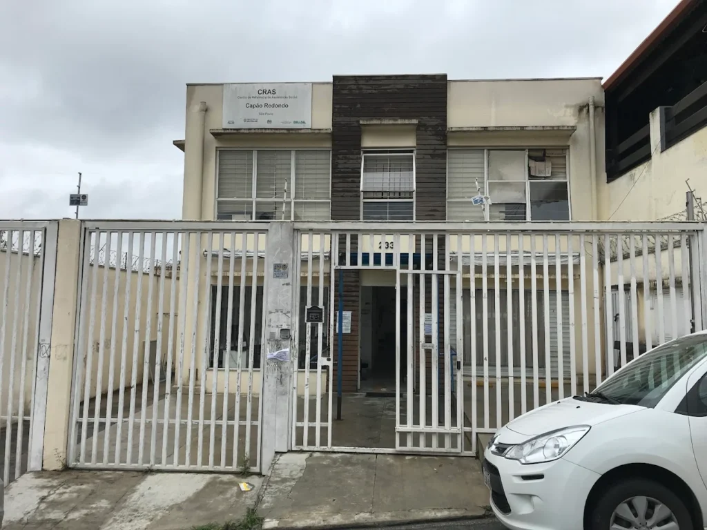 Fachada do CRAS Capão Redondo com portão metálico, prédio de dois andares e carro estacionado em frente à unidade.