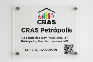 Placa de vidro montada na parede com o logotipo colorido do CRAS e os dizeres CRAS Petrópolis, Rua Frederico Boy Prussiano, 137 – Petrópolis, Belo Horizonte – MG, telefone (31) 3277-9578 e QR Code para acesso à página da unidade.